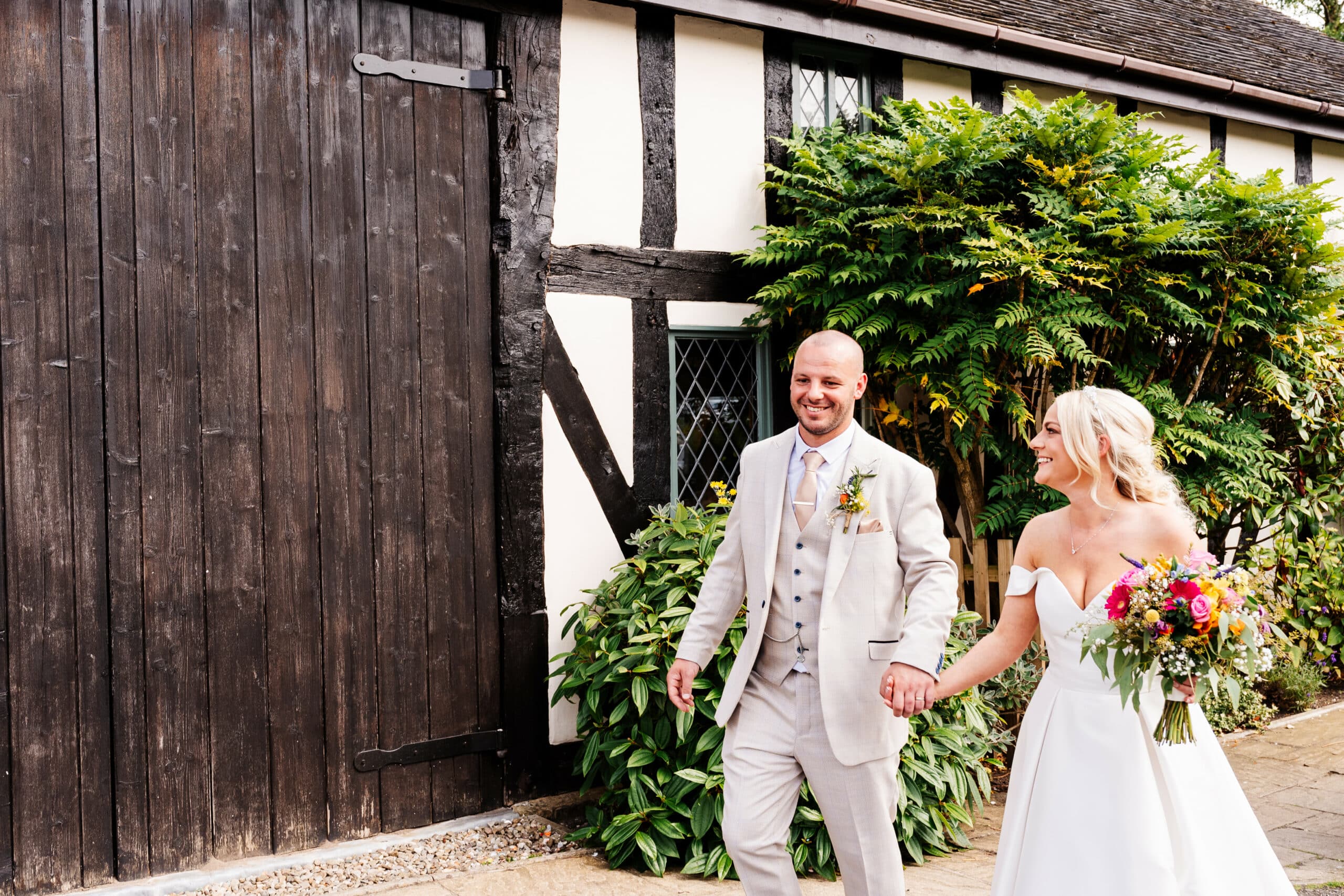A bride and groom walking hand in hand beside the barn at The Plough
